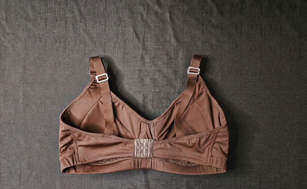 A brown structured underwire bra with adjustable shoulder straps and a lace-up center front, laid flat on a dark textured fabric background.