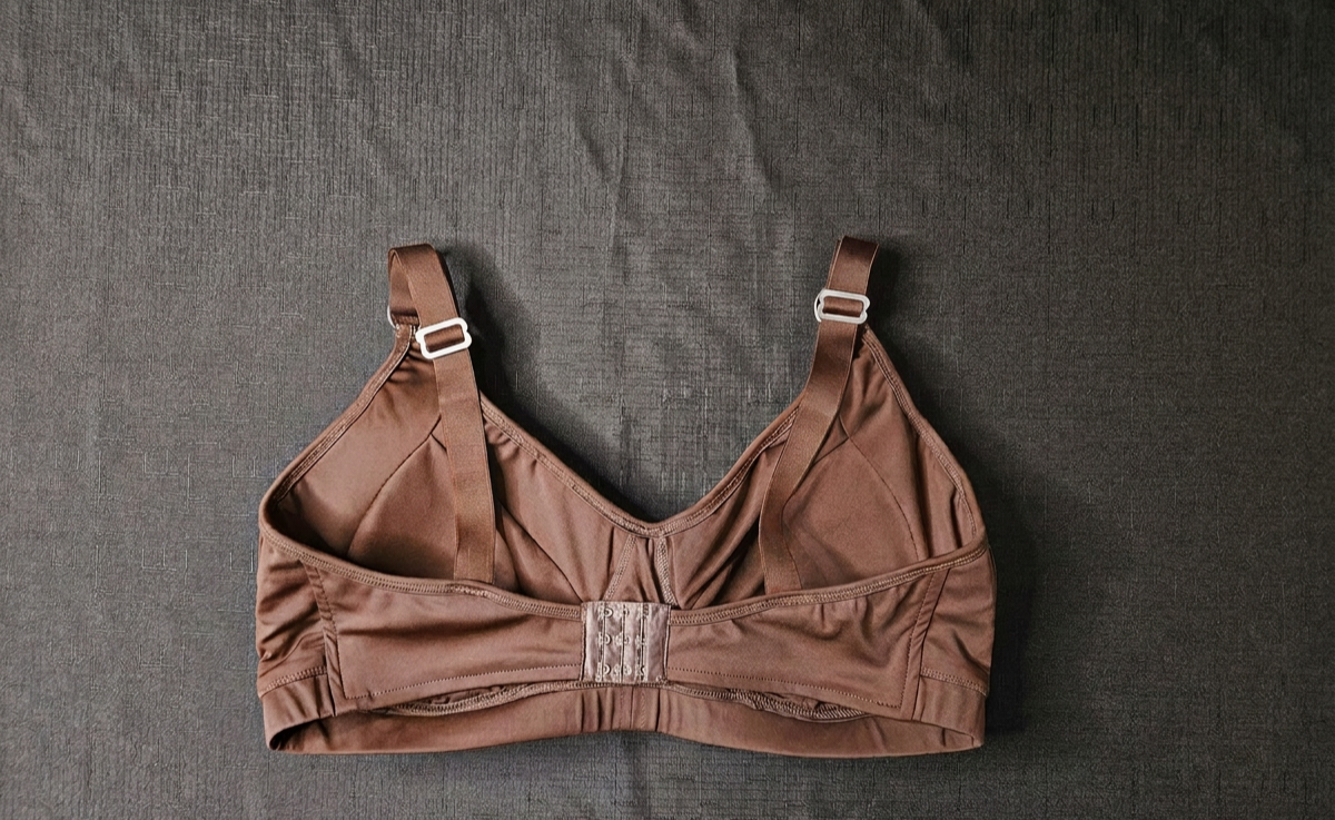 A brown structured underwire bra with adjustable shoulder straps and a lace-up center front, laid flat on a dark textured fabric background.