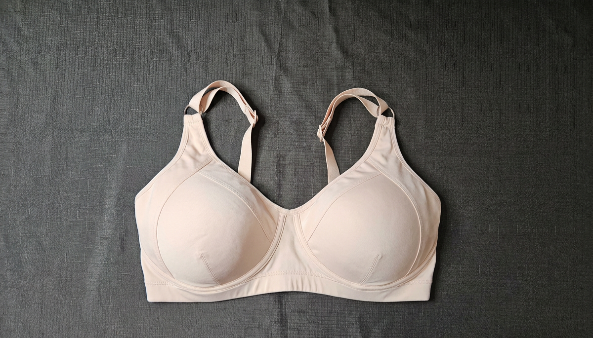 A light beige wireless bra with wide shoulder straps, laid flat on a textured dark gray fabric background.