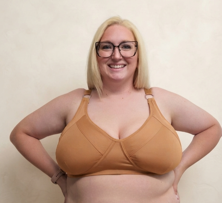 A smiling blonde woman wearing a tan molded-cup bra with shoulder straps, standing against a plain beige wall.