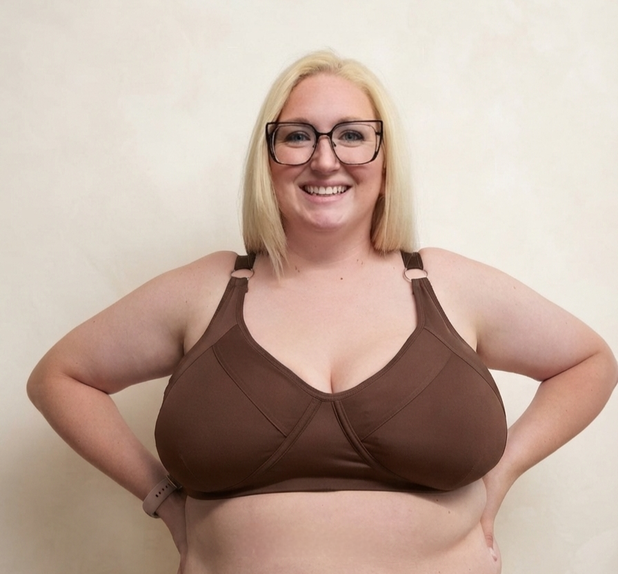 A smiling blonde woman wearing black-rimmed glasses and a brown bra, standing against a plain beige wall with her hands on her hips.