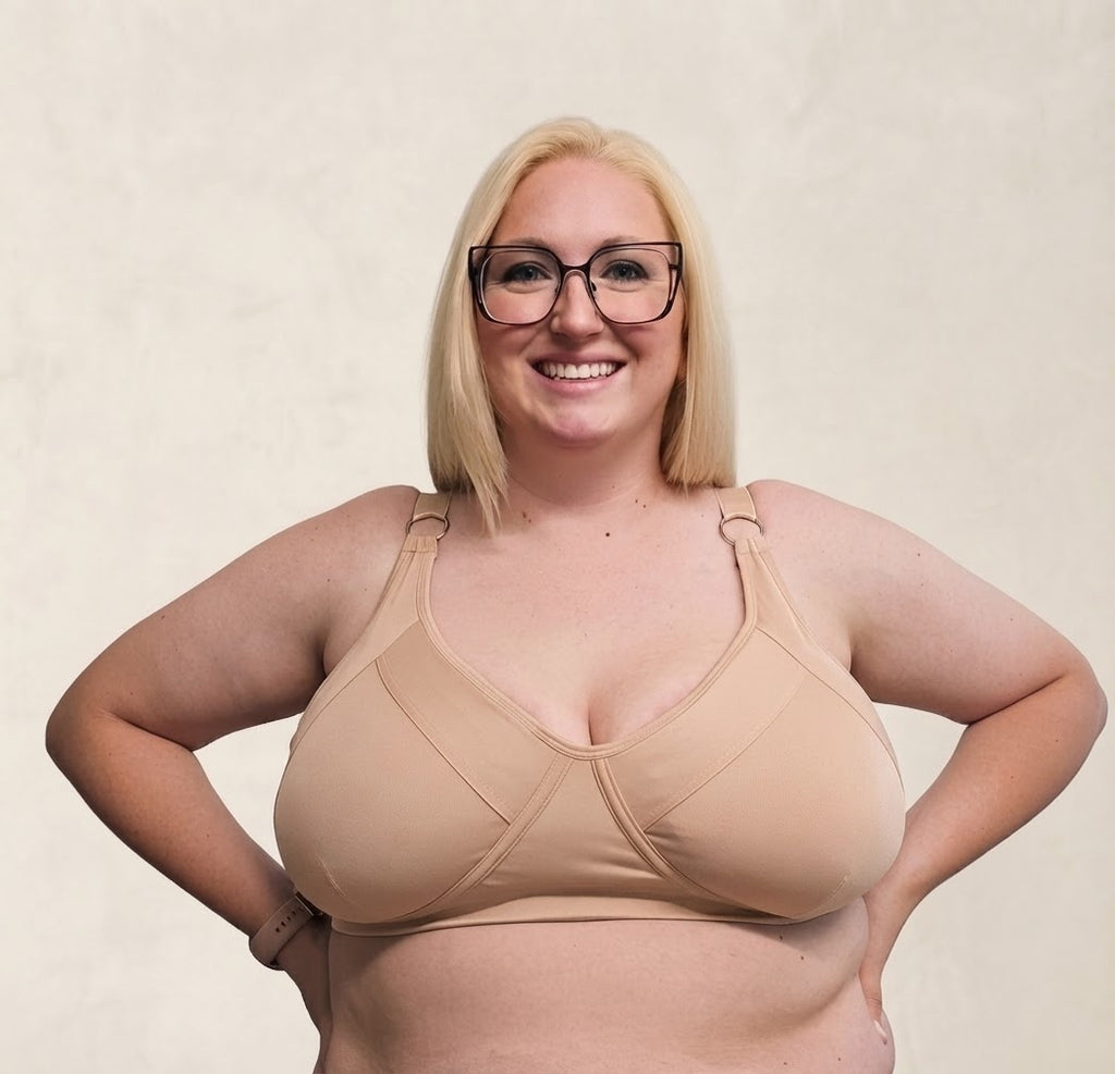 A smiling blonde woman wearing a beige underwire bra and glasses, posing with her hands on her hips against a textured beige background.