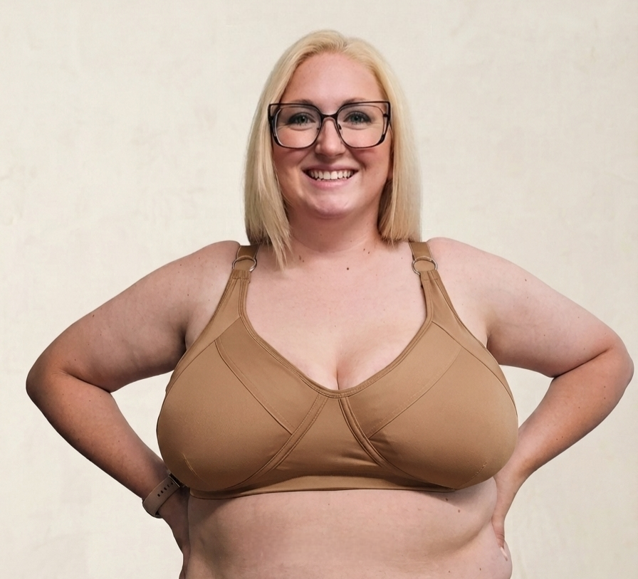 A smiling blonde woman wearing black-rimmed glasses and a tan supportive bra, posing with hands on hips against a light beige textured wall.